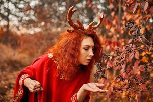 Woman in long red dress with deer horns in autumn forest.