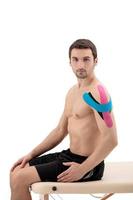 Male shoulder with physio tape, isolated on a white background. Male model sitting on a portable massage table