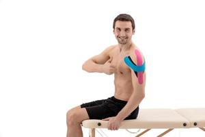 Male shoulder with physio tape, isolated on a white background. Male model sitting on a portable massage table