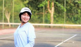 Hermosa mujer asiática con pelo corto, con sombrero y una amplia sonrisa en la cancha de tenis foto