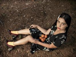 A beautiful girl of Caucasian appearance is sitting on the ground in a long dress, holding in it a lot of mandarin. view from above photo