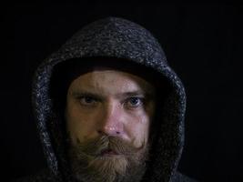 Retrato de un hombre con barba y bigote en el capó con un rostro serio sobre un fondo negro foto