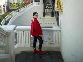 Atractiva chica rubia hipster con un sombrero negro tejido y chaqueta roja está de pie en las escaleras debajo del puente foto
