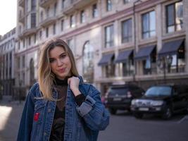 Chica atractiva con un cabello precioso en una chaqueta de mezclilla con el telón de fondo de un edificio moderno foto