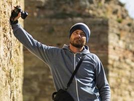 a bearded man wearing a knitted hat stands against a wall in a fortress with a camera in his hand and a camera bag over his shoulder photo