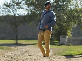 Un hombre con un suéter con capucha y gorro de punto está de pie sobre un fondo de árboles verdes con un palo y sonriendo foto