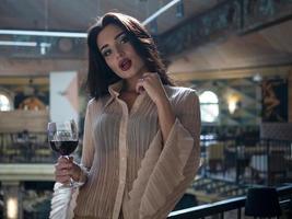 Hermosa chica modelo posando de pie con una copa de vino tinto en la mano en un restaurante