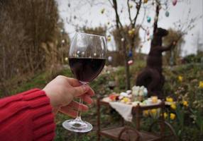 Copa de vino tinto en la mano femenina y árbol de Pascua con decoración y brunch de desayuno festivo en la mesa móvil retro en el jardín. foto