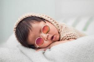 adorable bebé recién nacido durmiendo pacíficamente sobre una manta blanca. el lleva gafas de sol foto