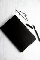 Pendientes cuaderno de cuero negro y bolígrafo sobre mesa de madera blanca, conjunto de negocios y joyas foto