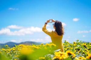 mujer feliz verano despreocupado en campo de girasol en primavera. Alegre mujer asiática multirracial manos formando una forma de corazón en el campo de girasoles foto