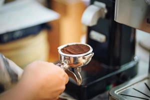 Hand of barista holding portafilter filled with ground coffee preparing to brew with machine. Selective focus