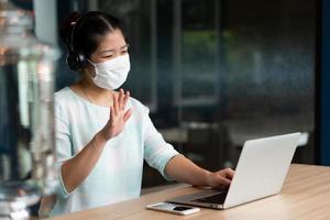 Portrait of young asian woman wearing face mask and headphones and working remotely by using computer video call confernece at coworking space. social distancing and new normal lifestyle concept photo