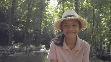 portret schattig meisje met strohoed lacht en kijkt naar videocamera terwijl ze tijdens de zomervakantie in tropisch bos verblijft. Thailand. video