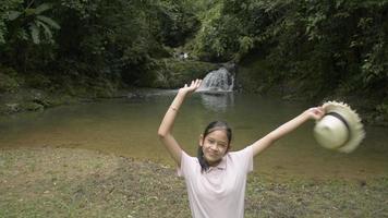 Cheerful pretty girl raise arms to the air with straw hat and looking at camera. Female teenager standing alone near small waterfall in tropical forest. Aerial drone shot. video