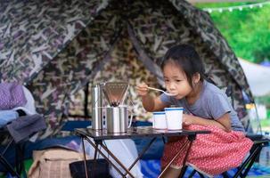 Little Asian girl sitting and eating breakfast in front of tent while camping photo