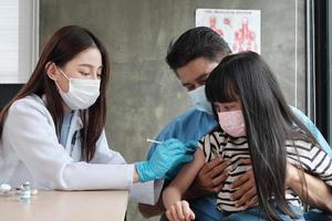 Female doctor with face mask vaccinating Asian girl to prevent coronavirus COVID-19 At the pediatrics clinic in kid hospital with father nearby. Injections treat illnesses, cause pain in children. photo