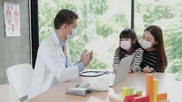 A mother with a face mask brings her sick daughter to an exam appointment with an Asian male doctor at the pediatrics department of a kid's clinic. To treat illnesses and to consult on family health. video