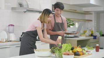 Happy Asian beautiful young family couple husband and wife cooking food vegetable salad in kitchen together at home. The man and woman according online cooking class to recipe on laptop computer video