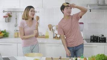 Happy Asian beautiful family couple husband and wife singing in kitchenware microphones in kitchen together having fun dance listen music at home. Two people dancing slow motion