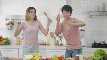 Happy Asian beautiful family couple husband and wife singing in kitchenware microphones in kitchen together having fun dance listen music at home. Two people dancing slow motion video