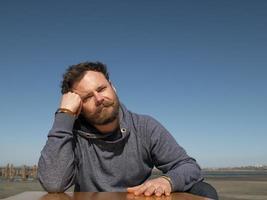 pensive man with a beard sitting at a coffee table against a blue sky photo