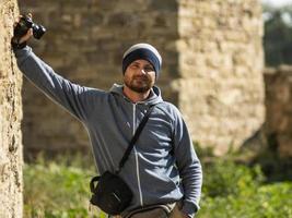 a bearded man stands against a wall in a fortress with a camera photo