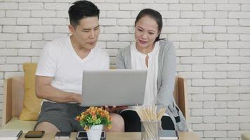 Happy Asian family couple husband and wife work and using laptop computer analyzing their finances discuss. Cheerful loving couple smile shopping order products online together from home by computer video