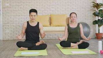 gelukkige Aziatische mooie levensstijl familie paar doet yoga zittend mediteren op lotus pose training thuis samen op mat met gesloten ogen. twee mensen sporten gezond concept. slow motion video