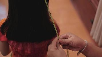 Close-up VDO. Cute long, black hair Asian girl is being braided in multi-colored threads by woman's hand in beauty salon. Child fashion, weaving pigtails with decoration yarn or knitting wool. video