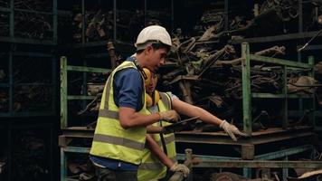 trabajadores asiáticos masculinos y femeninos en uniformes y cascos, control de existencias de empleados de la fábrica de almacén de repuestos de automóviles. usando una tableta para verificar motores viejos y pedidos de máquinas para la industria automotriz. video