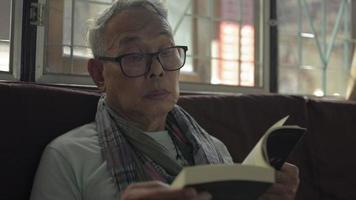 Senior hombre de cabello gris con anteojos leyendo un libro de novelas en casa. video