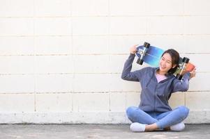 Asian woman with surfskate sitting against concrete wall photo
