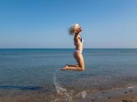 Blond girl in bikini jumping up with bent legs, splashes of water photo