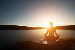 silueta de mujer sana está practicando yoga lago durante la puesta de sol. foto