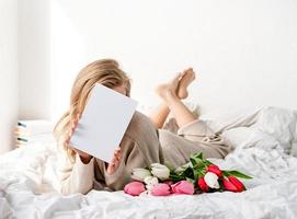 Mujer feliz acostada en la cama sosteniendo flores de tulipán y tarjeta en blanco foto