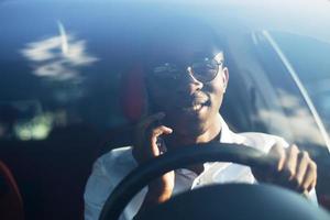 Feliz afroamericano conduciendo un coche con un teléfono, en el verano