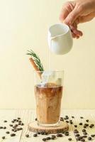 Pouring milk in black coffee glass with ice cube, cinnamon, and rosemary on wood background photo