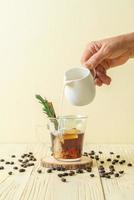 Verter la leche en un vaso de café negro con cubitos de hielo, canela y romero sobre fondo de madera