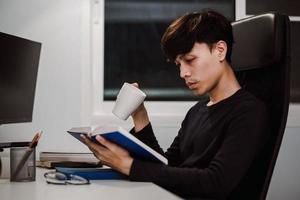 Young handsome asian man reading book at work desk late at night photo