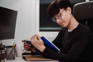 Joven apuesto hombre asiático leyendo un libro en la mesa de trabajo a altas horas de la noche foto