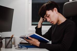 Young handsome asian man reading book at work desk late at night photo