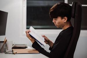 Young handsome asian man reading book at work desk late at night photo