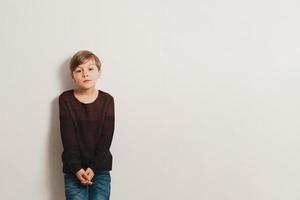 A cute boy with unhappy face, leans to a white wall photo