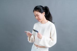 Retrato de una joven asiática feliz mostrando una tarjeta de crédito de plástico mientras sostiene el teléfono móvil sobre fondo gris foto