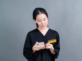 Portrait of a happy young Asian girl showing plastic credit card while holding mobile phone on grey background photo