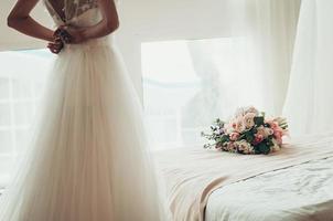 wedding bouquet on a bed, blurred bride buttoning her dress, back view photo