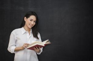 young asian woman reading book while standing against blackboard photo
