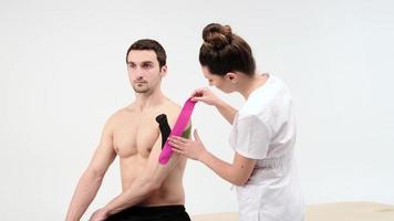 The doctor glues a special treatment tape to mail shoulder. Physiotherapist sticks kinesio tapes to the shoulder of patient, kinesiology taping, kinesiological therapy, athlete are recovering after injury of loins, 4k photo