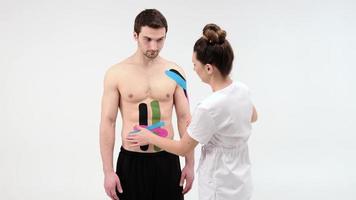 Terapeuta de sexo femenino que aplica la cinta de la kinesiología en el abdomen de un hombre en el fondo blanco. Mujer prepara paciente masculino para pegar cinta adhesiva kinesio foto